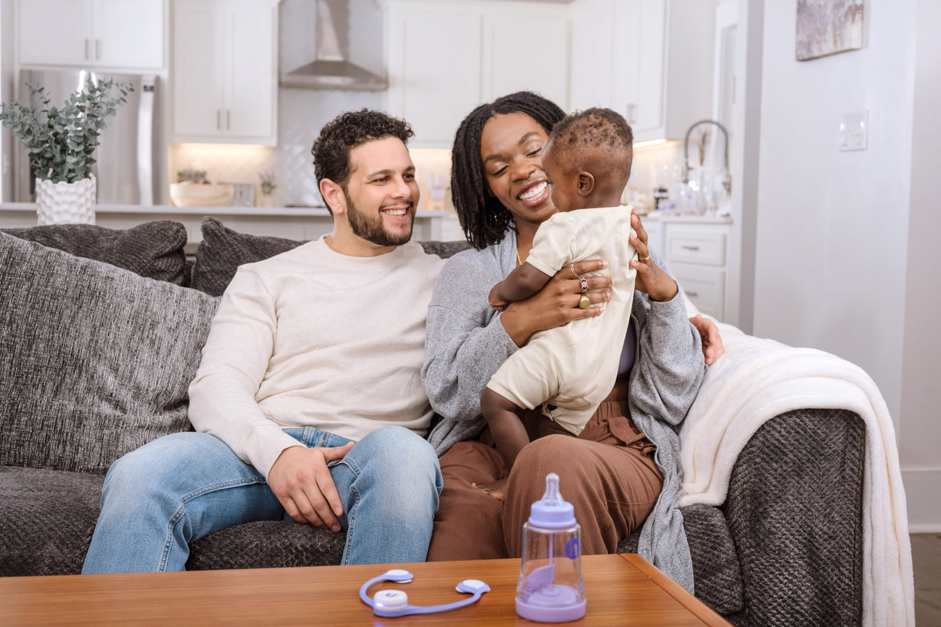 Family of three sitting on a couch in a living room with a baby bottle and toy on the coffee table.
